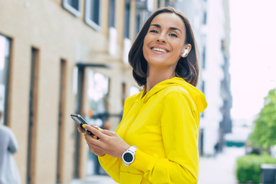 Sárga felsős, vállig érő, barna hajú, mosolygós, fiatal nő mobiltelefonozik.